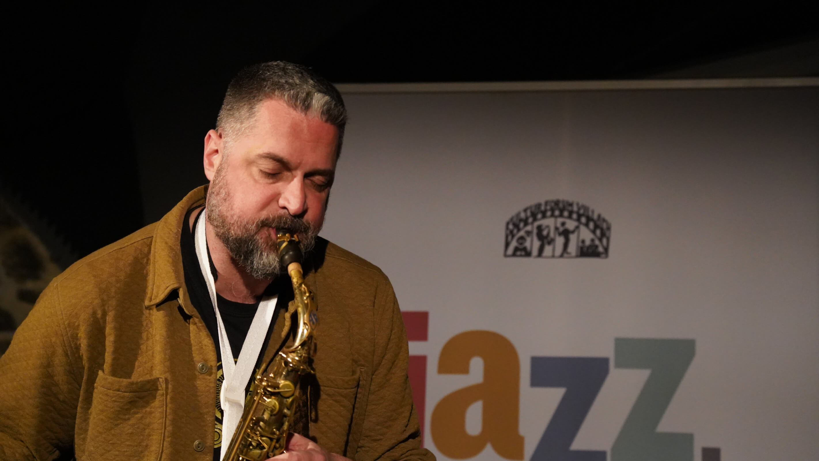 Altsaxophonist in brauner Jacke spielt vor Jazz-Villach-Banner.