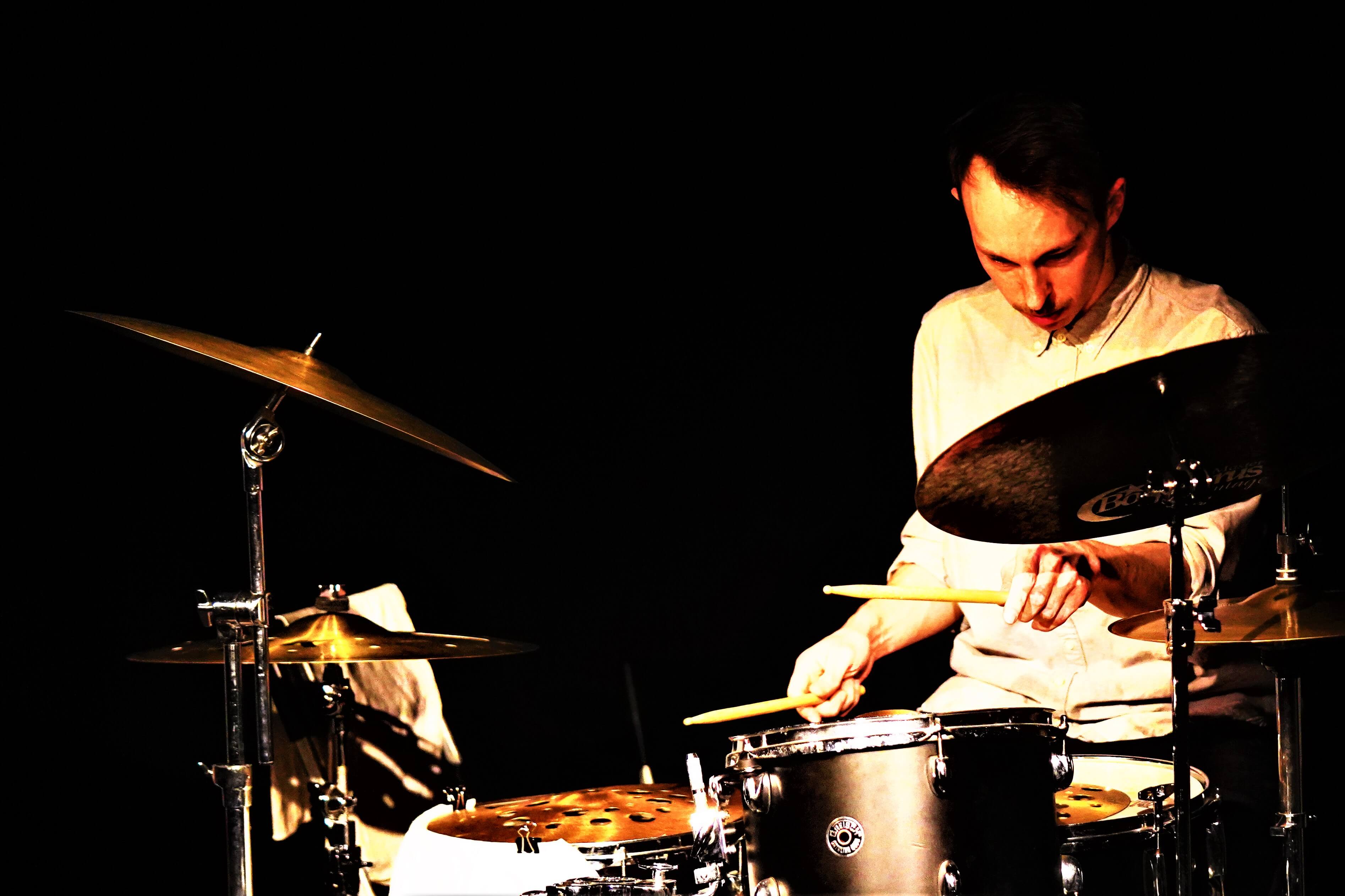 Schlagzeuger, eingerahmt von Becken und Toms vor schwarzem Hintergrund.