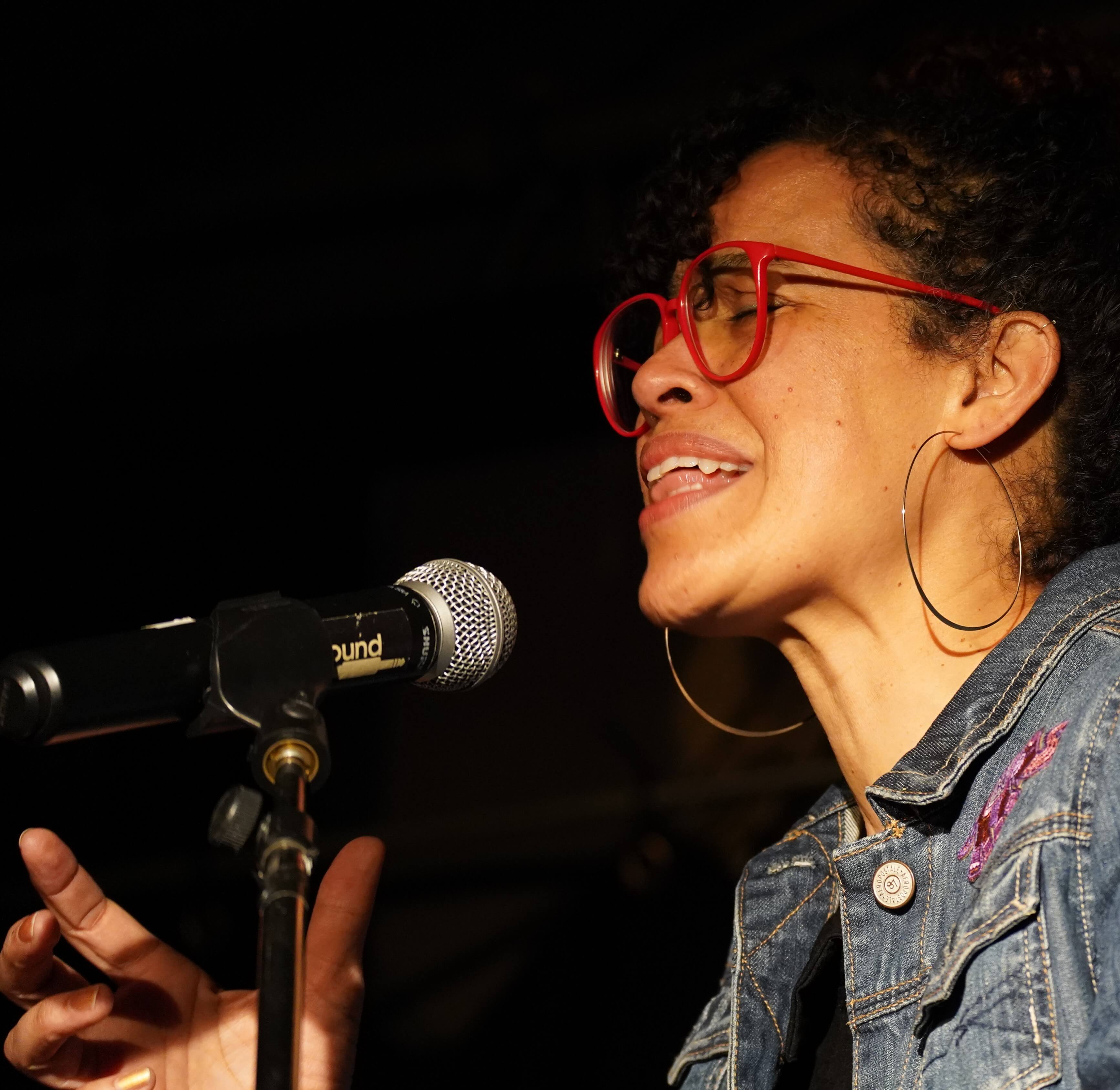 Sängerin in Jeansjacke und roter Brille am Mikrofon, gestikuliert mit einer Hand.