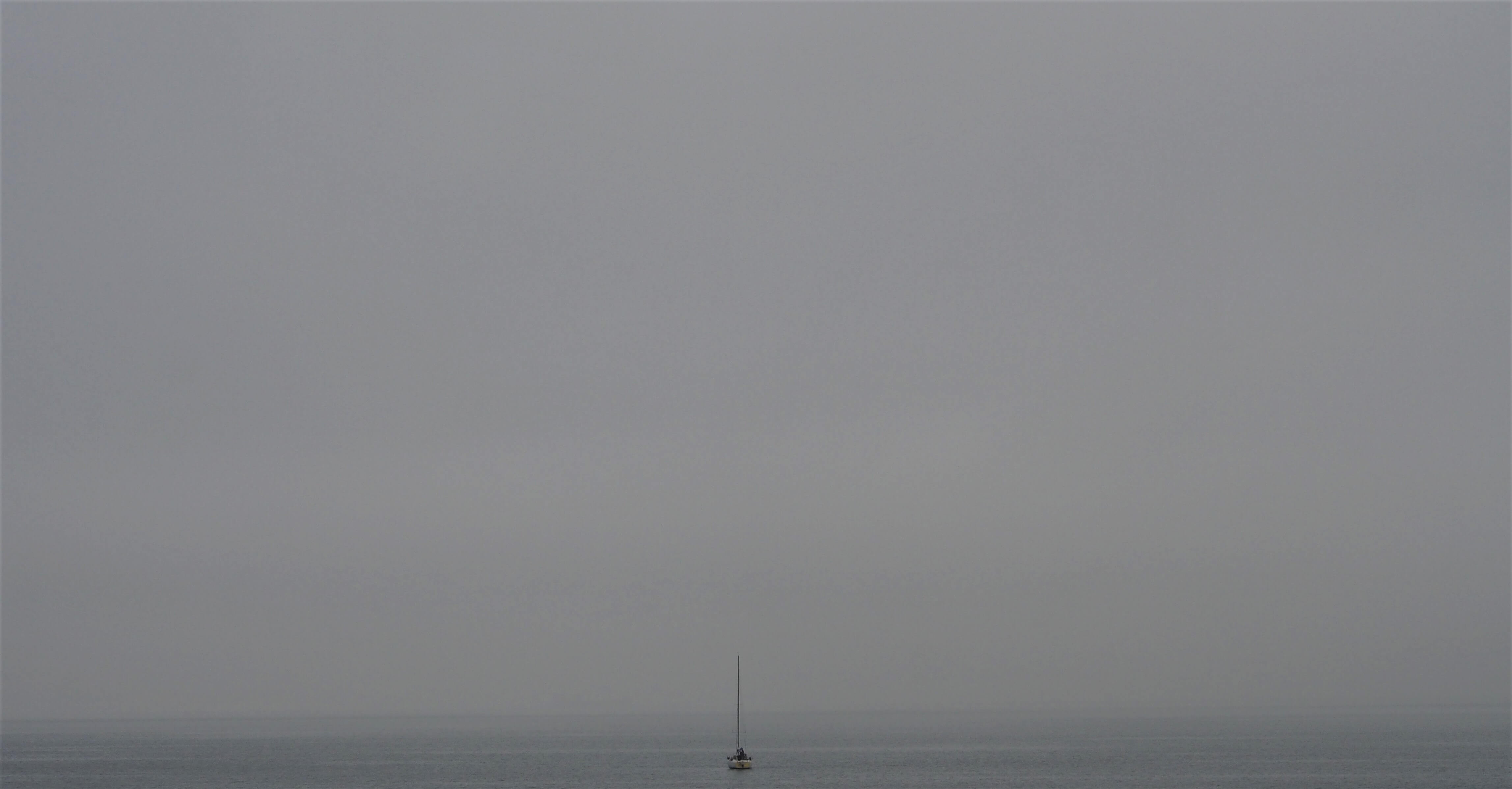 Minimalistische Meereslandschaft; ein einsames Segelboot auf ruhigem Wasser unter einem weiten, nebligen Himmel.