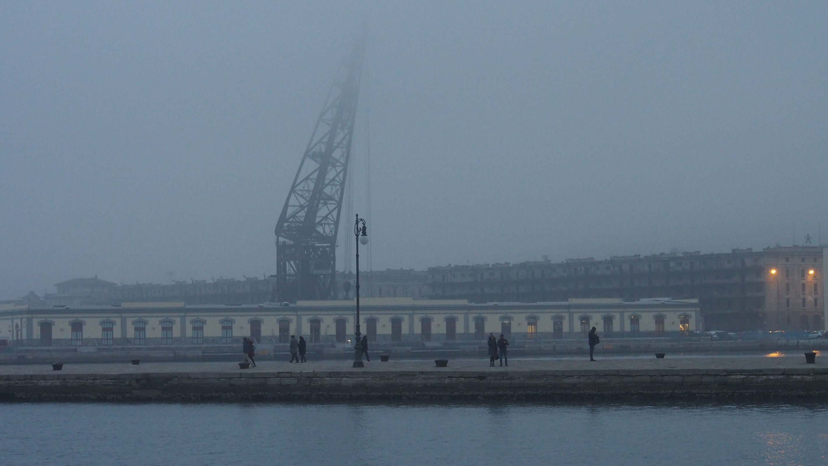 Alte Hafengebäude, ein hoher Kran und Menschen erscheinen als verblassende Silhouetten im Hafennebel.