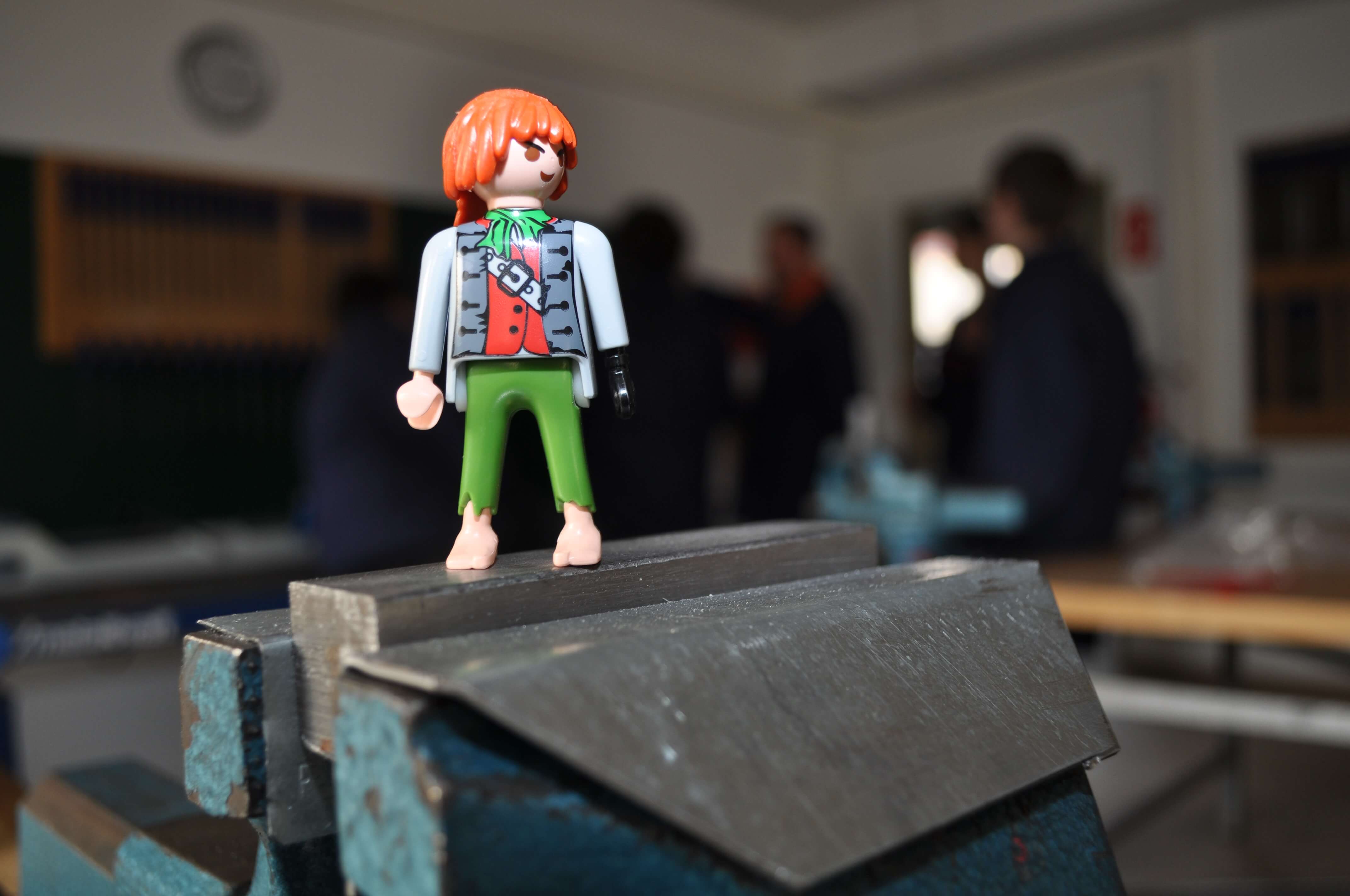 Playmobil-Piratenfigur steht auf einem Schraubstock in einer Werkstatt mit unscharfen Personen im Hintergrund.