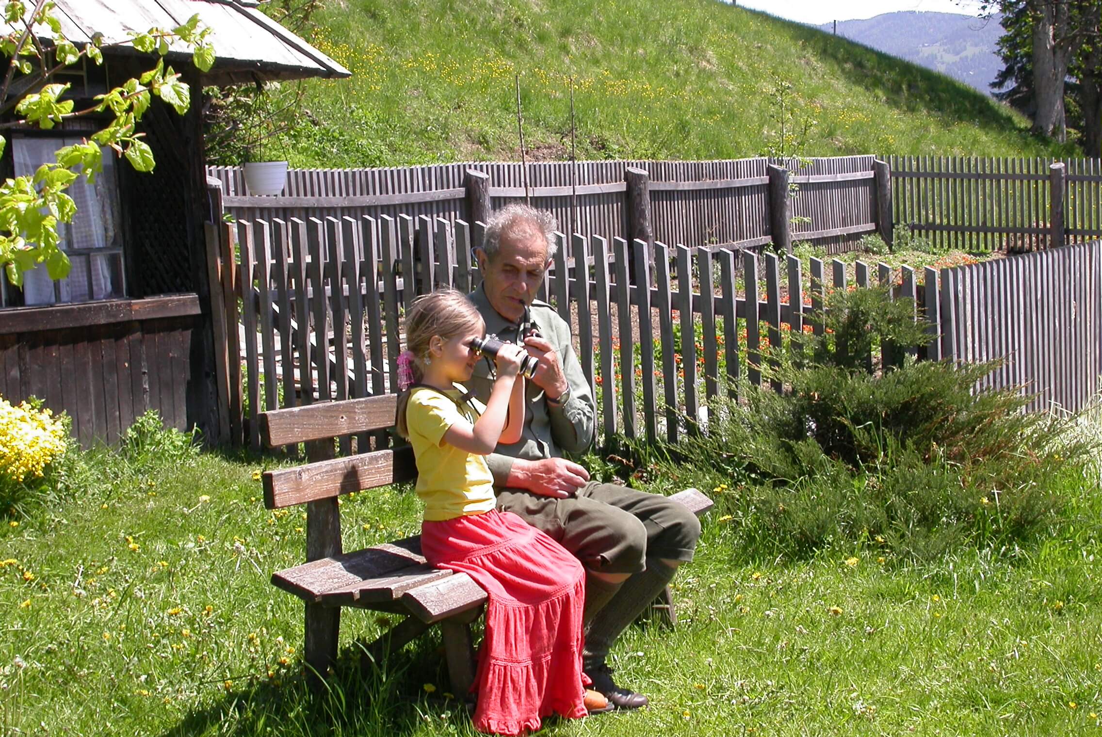 Mein Vater auf einer einfachen Holzbank im Garten, während ich als kleines Kind neben ihm sitze und sein Fernglas vor die Augen halte.